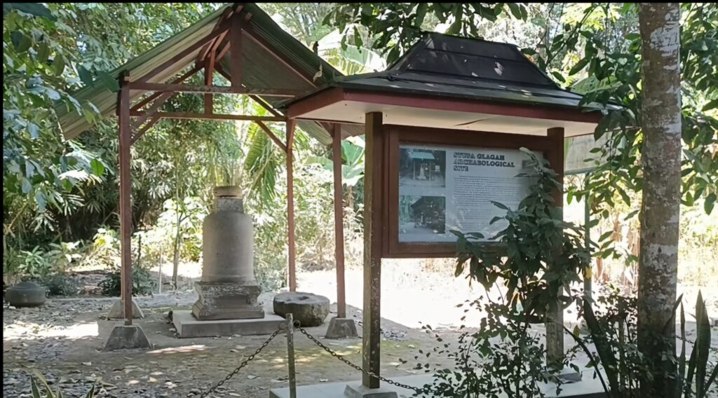 jjjh2 - Mabur.co Outdoor garden shrine with a wooden shelter and a glass-front information board, surrounded by trees and stone bases.