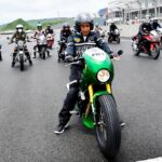 Motorcyclist wearing a helmet on a bright green sport bike leads a line of riders on a race track, with spectators in the background.