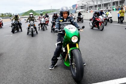 Motorcyclist wearing a helmet on a bright green sport bike leads a line of riders on a race track, with spectators in the background.
