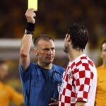 Soccer referee raises a yellow card to a player during a match, with a Croatian team member in a red-and-white checkered jersey nearby.