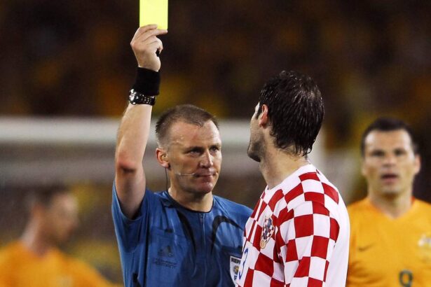 Soccer referee raises a yellow card to a player during a match, with a Croatian team member in a red-and-white checkered jersey nearby.