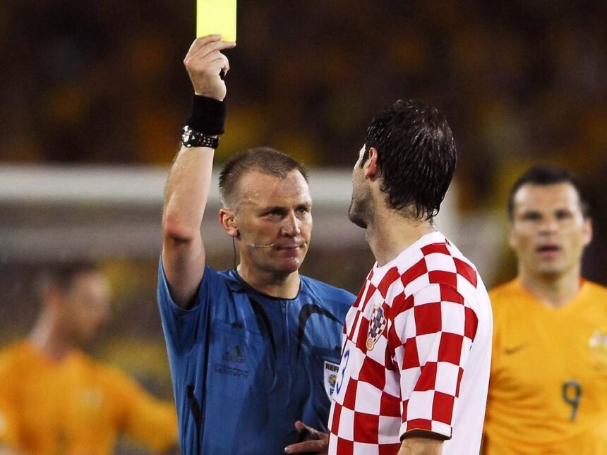 Soccer referee raises a yellow card to a player during a match, with a Croatian team member in a red-and-white checkered jersey nearby.
