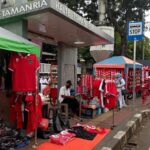 Outdoor street market with red jerseys and scarves on display along a sidewalk near a stop sign; Halte Taman Ria signage overhead.