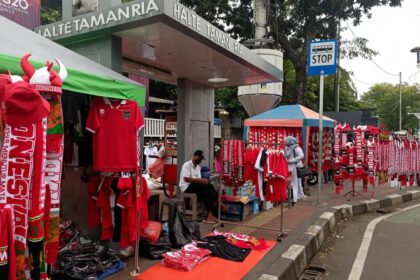 Outdoor street market with red jerseys and scarves on display along a sidewalk near a stop sign; Halte Taman Ria signage overhead.
