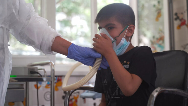 Boy sits in a chair while a clinician in full PPE holds an oxygen mask to his face, assisting his breathing.