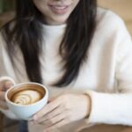 Person in a cream sweater holds a white cup with latte art, seated at a wooden table, smiling slightly.