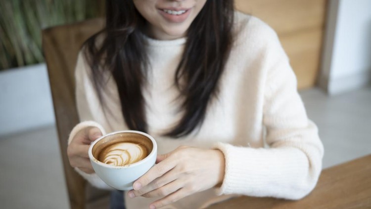 Person in a cream sweater holds a white cup with latte art, seated at a wooden table, smiling slightly.