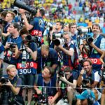 Group of photographers with large lenses and cameras gathered behind a barrier at a stadium event, many wearing blue vests and looking toward the crowd/scene nearby.