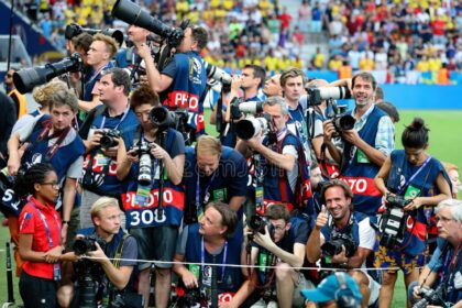 Group of photographers with large lenses and cameras gathered behind a barrier at a stadium event, many wearing blue vests and looking toward the crowd/scene nearby.