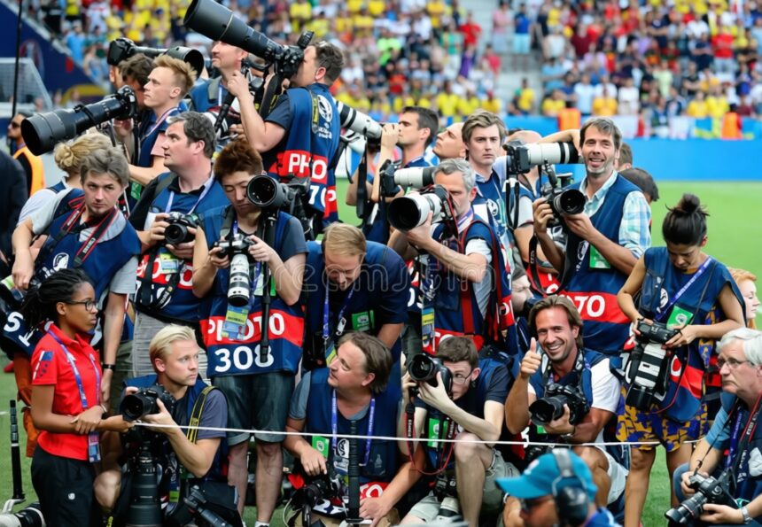 Group of photographers with large lenses and cameras gathered behind a barrier at a stadium event, many wearing blue vests and looking toward the crowd/scene nearby.