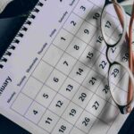 Close-up of a calendar page with a grid of dates, reading glasses resting on it and a spiral notebook in the background.