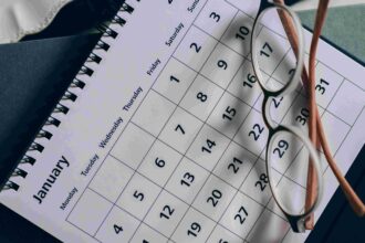 Close-up of a calendar page with a grid of dates, reading glasses resting on it and a spiral notebook in the background.
