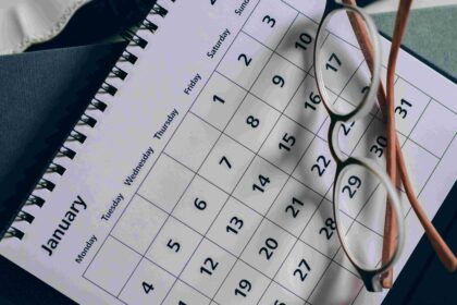 Close-up of a calendar page with a grid of dates, reading glasses resting on it and a spiral notebook in the background.