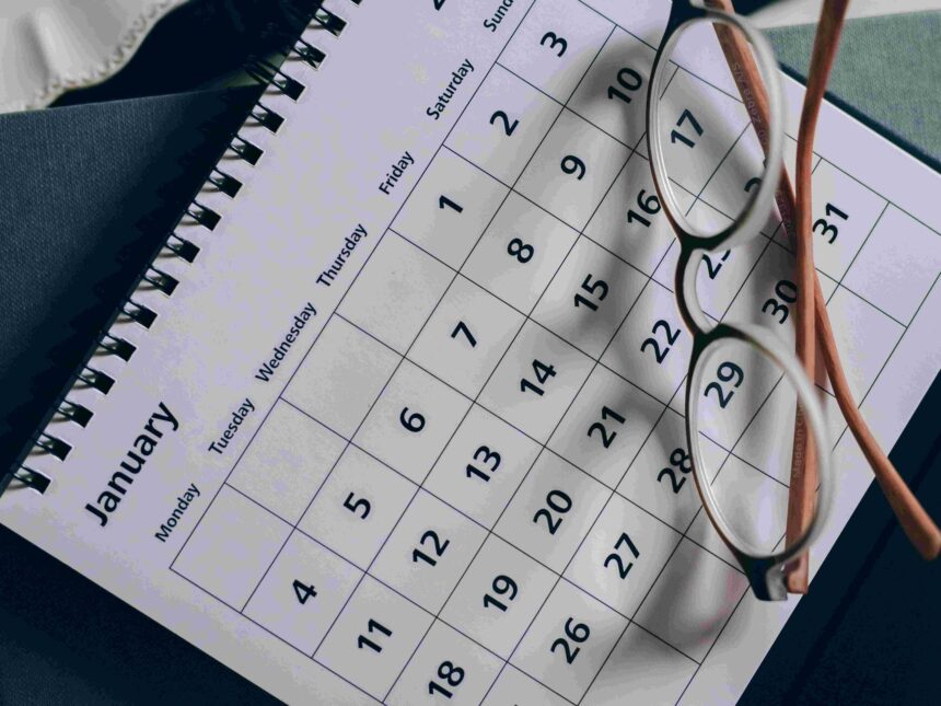 Close-up of a calendar page with a grid of dates, reading glasses resting on it and a spiral notebook in the background.