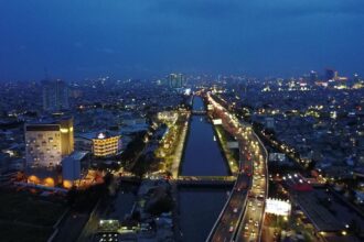 Aerial night view of a city with a central canal flanked by illuminated roads and dense, glowing buildings.