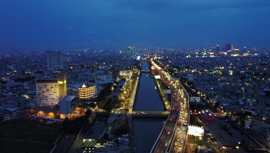Aerial night view of a city with a central canal flanked by illuminated roads and dense, glowing buildings.