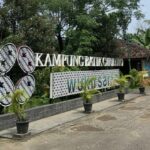 Signage for Kampung Batik Grolo with a decorative 'Welcome' panel and potted plants along a paved walkway.