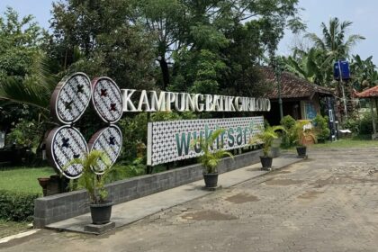 Signage for Kampung Batik Grolo with a decorative 'Welcome' panel and potted plants along a paved walkway.