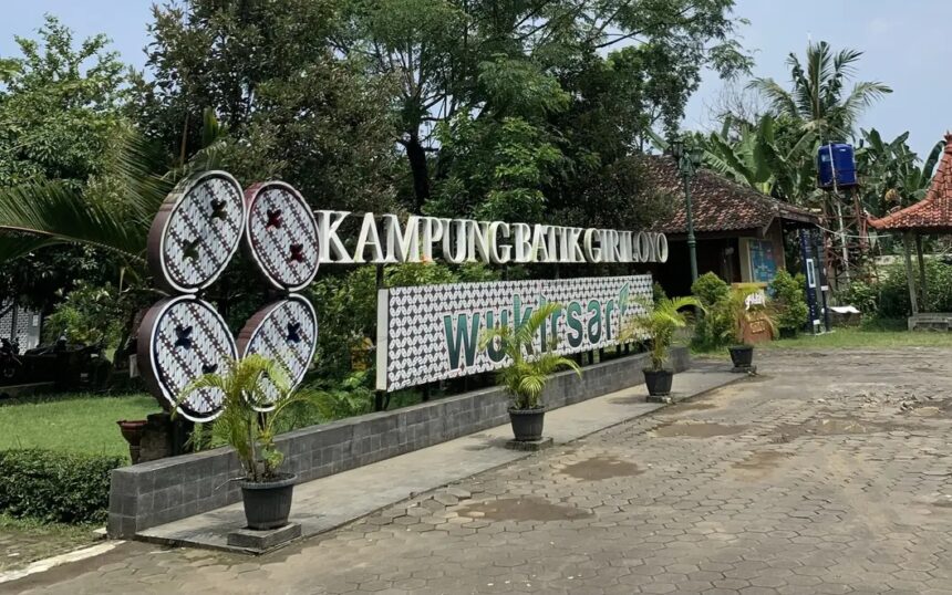 Signage for Kampung Batik Grolo with a decorative 'Welcome' panel and potted plants along a paved walkway.