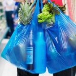 Person carrying two blue grocery bags filled with fresh produce and a bottle, in a supermarket aisle