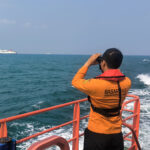 Person in an orange life jacket on a boat deck uses binoculars to scan the sea, with a distant ship on the horizon in calm blue waters.