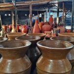 Outdoor pottery yard filled with large and small clay pots under a simple shelter; many unfinished vessels in earthy tones.