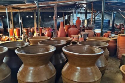 Outdoor pottery yard filled with large and small clay pots under a simple shelter; many unfinished vessels in earthy tones.