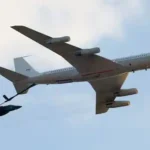 A large white airliner in flight towing a smaller black fighter jet behind it by a tether.