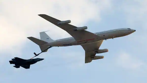 A large white airliner in flight towing a smaller black fighter jet behind it by a tether.