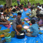 Large group of people seated on a blue tarp outdoors, sharing a communal meal and eating rice from metal bowls under trees.