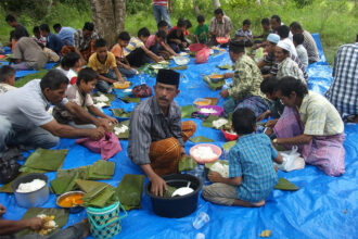 Large group of people seated on a blue tarp outdoors, sharing a communal meal and eating rice from metal bowls under trees.
