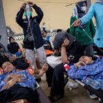 Children lying on medical stretchers, resting under blankets as caregivers and volunteers attend in a crowded relief clinic.