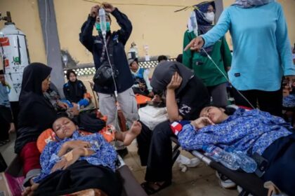 Children lying on medical stretchers, resting under blankets as caregivers and volunteers attend in a crowded relief clinic.