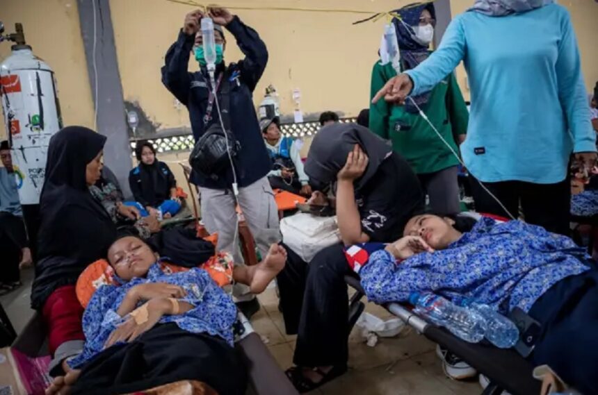 Children lying on medical stretchers, resting under blankets as caregivers and volunteers attend in a crowded relief clinic.