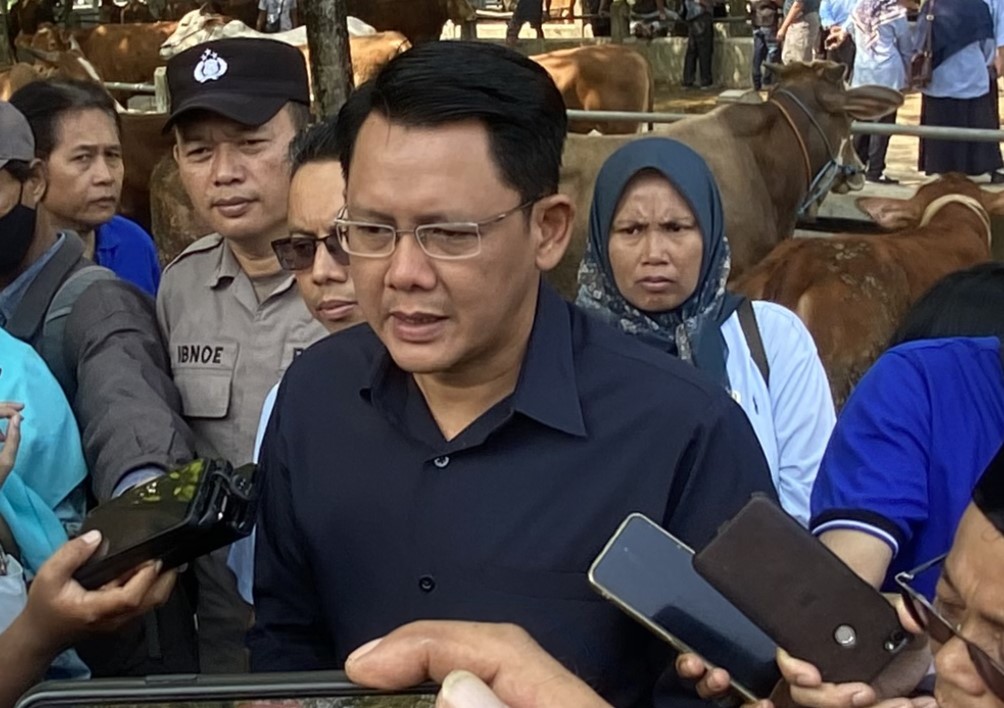 Man in a dark shirt speaks to reporters as cameras and smartphones are aimed at him, with a crowd behind in an outdoor setting.