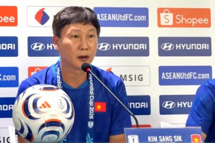 Asian man in a blue shirt speaks at a press conference with a soccer ball on the table and sponsor logos in the background, including Hyundai and Shopee.