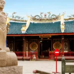 Stone statue of a warrior in front of a Chinese temple with dragon-adorned roof.