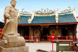 Stone statue of a warrior in front of a Chinese temple with dragon-adorned roof.