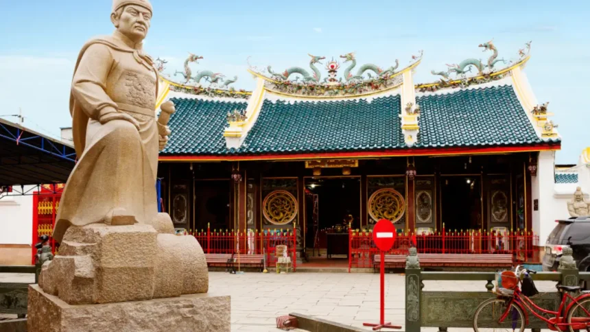 klenteng-tay-kak-sie - Mabur.co Stone statue of a warrior in front of a Chinese temple with dragon-adorned roof.