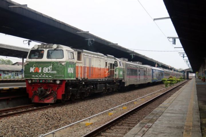 Diesel locomotive in green, white, and orange pulling a passenger train at a platform station with a shelter overhead.