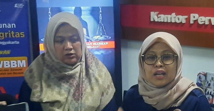Two women wearing hijabs stand indoors near blue and red banners; one woman speaks while the other looks down.