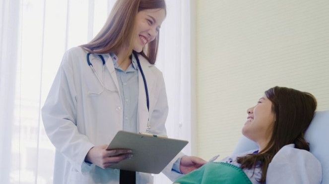 Female doctor in a white coat with a stethoscope and clipboard talking to a patient in a hospital bed.