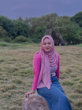 krw - Mabur.co Woman in a pink hijab and top sits on a log in a grassy field with trees in the background.