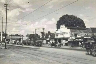 Black-and-white street scene with vintage cars, utility poles, and storefronts; a horse-drawn cart on the right.