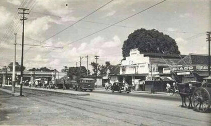 Black-and-white street scene with vintage cars, utility poles, and storefronts; a horse-drawn cart on the right.