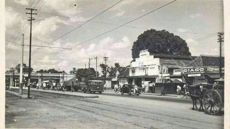 Black-and-white street scene with vintage cars, utility poles, and storefronts; a horse-drawn cart on the right.