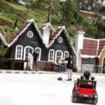 Row of chalet-style houses with white trim along a street; people chat and sit outside while a child drives a red toy car in the foreground.
