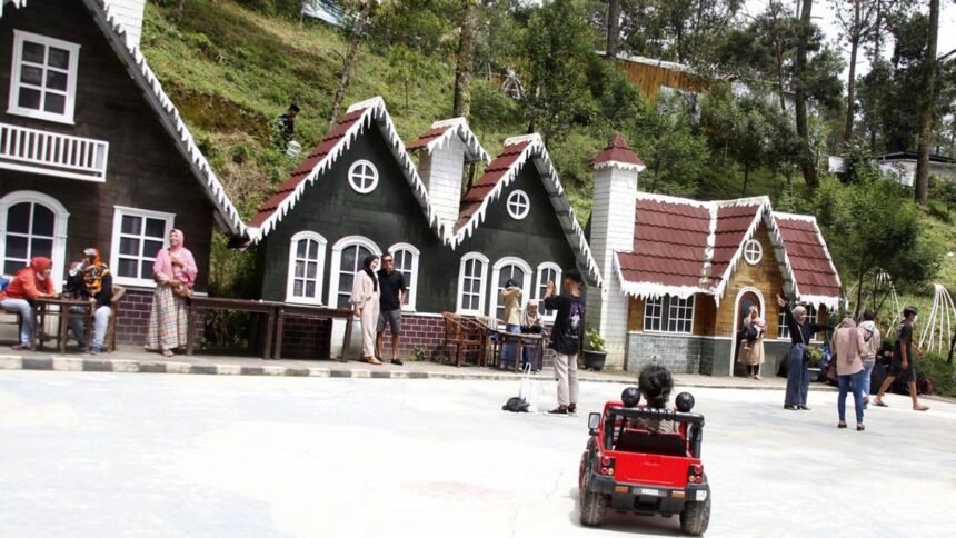 Row of chalet-style houses with white trim along a street; people chat and sit outside while a child drives a red toy car in the foreground.