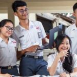 Five students in matching uniforms pose and study together, smiling around a table with notebooks and a laptop in a classroom or library setting.