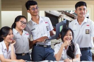 Five students in matching uniforms pose and study together, smiling around a table with notebooks and a laptop in a classroom or library setting.